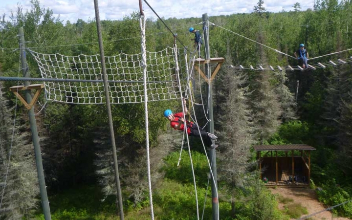 On a high ropes course, three people wearing safety gear and secured by ropes make their way through the course. 